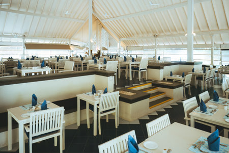 Wide angle shot of a restaurant with white furniture, blue napkins, a black floor, and a pyramidal ceiling. It has two levels, and steps join them, creating an open atmosphere and luxurious dining space.の写真素材