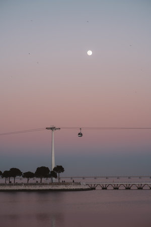 A low-key, vertical view of a beautiful scene featuring a full moon, pink sky sunset, the Tagus river, and a cable car. The overall effect is a serene and dreamy magical scene captured in Lisbonの写真素材
