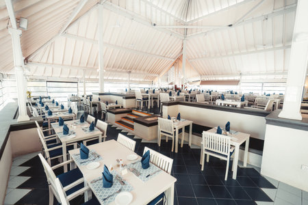 Wide-angle shot of a serene Maldivian restaurant divided into two levels, with white furniture, blue napkins and cushions, a black floor, and an open and airy ambiance, very invitingの写真素材