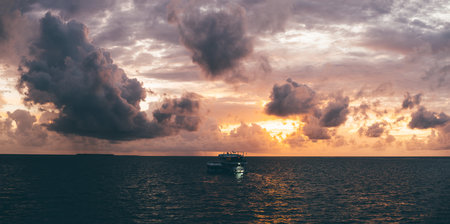 A stunning sunset low-key panoramic view of a boat in the sea. The orange sun is partially hidden behind violet and pink clouds; The boat's silhouette is highlighted against the dramatic backdropの写真素材