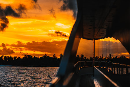 A low-key shot of a sunset in the Maldives, from a yacht sailing on the sea. A breath-taking sunset, with some visible clouds. The shot faces towards land, where trees can be seen in the distance.の写真素材