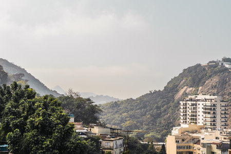View from an apartment in Rio de Janeiro, surrounded by lush mountains covered in green trees, humble buildings dot the landscape; The sunny day is accompanied by light fog cloudsの写真素材