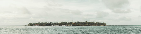 Maldives; Panorama of a Maldivian islet featuring lush green trees, surrounded by the vast calm blue sea, gray clouds in the sky contrasting with the overall tranquil sceneの写真素材
