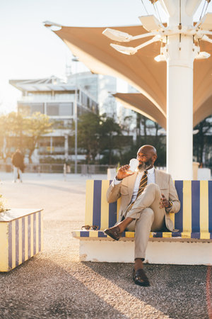 Lisbon; A sophisticated and bald black guy sits on a colorful outdoor bench while taking a sip of coffee from his takeaway cup, he has his legs crossed revealing his ankles, on a sunny afternoonの写真素材