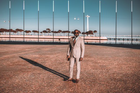 A stylish black man in a light-colored suit and striped tie stands on a brown cobblestone floor, with flag poles in the background and a glimpse of the river, under a beautiful blue skyの写真素材