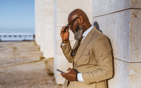Outdoors, a thoughtful African businessman stands by a limestone pillar. Holding his cell phone and adjusting the sunglasses on his face embracing the sunny day with the blue river as a backdropの写真素材