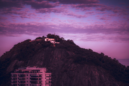 Long-focus shot of an evening mountain in the dusk with a fort on the top and the roof of a residential building in the foreground in toxic modern pink and purple colors with a partly cloudy sky aboveの写真素材