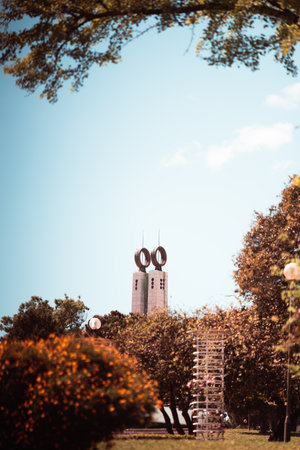 A vertical medium telephoto shot of a beautiful park in warm summer and autumn colors with selective focus on the tower in the background with two rings of a Monumento 25 de Abril in Lisbon, Portugalの素材 [FY310209757342]