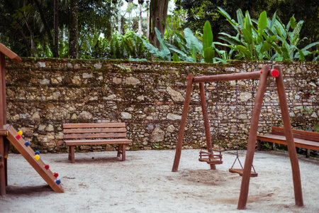 Quiet children's playground nestled in a tropical park, featuring a wooden bench, swing set, and colorful climbing structure. Lush foliage and a historic stone wall create a serene backdropの写真素材