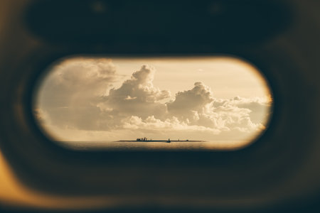 View of a small island in the distance through an oval porthole, with dramatic clouds in the sky and calm ocean waters. Perfect for travel, adventure, and seascape themesの写真素材