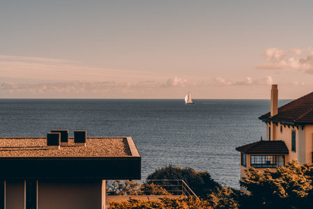 Scenic coastal view with a sailboat on the horizon, framed by residential buildings and greenery. The calm ocean and pastel sky at sunset create a tranquil atmosphere. Ideal for travel themesの写真素材
