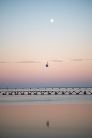 A vertical scenic view of a cable car traveling over a calm body of water with a bridge in the background, captured during a serene sunset. The sky transitions from pink to blue with the moon visibleの写真素材