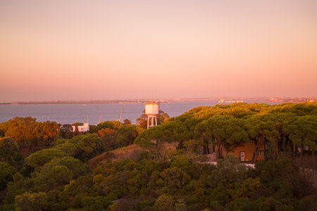 A scenic view of Almada, Portugal, at sunset featuring a water tower surrounded by lush greenery with a backdrop of the sea and distant cityscape under a dramatic pastel pink skyの写真素材