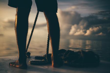 Tilt-shift low-key shot of a person standing barefoot on a boat deck, holding a rope. The blurred background features a calm sea and dramatic clouds at sunset, creating a serene atmosphereの写真素材