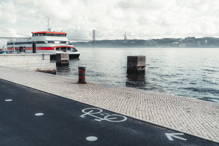 Modern ferry docked at a pier in Lisbon with the 25 de Abril Bridge and Cristo Rei statue visible in the background. Bicycle lane in the foreground adds an urban touch to the scenic waterfront viewの写真素材
