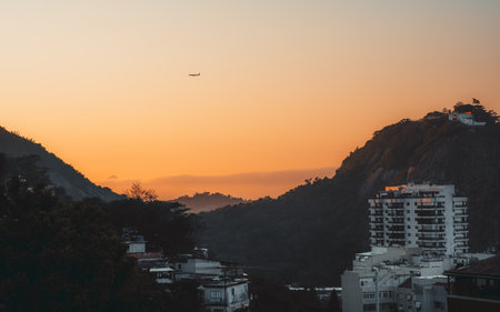 Scenic view of Rio de Janeiro at sunset or sunrise with a plane flying in the orange sky. Hills and mountains surround the city buildings, creating a picturesque and dramatic landscapeの写真素材