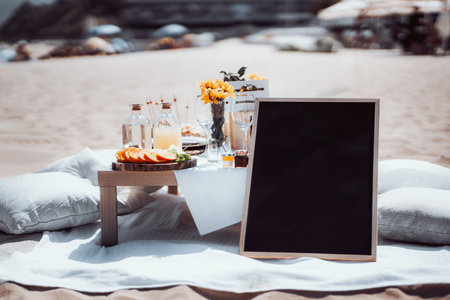 Elegant beach picnic setup with a wooden table featuring sliced citrus fruits, glass bottles, and a bouquet of yellow flowers. Blank chalkboard for text. Background includes sand and beach umbrellasの写真素材