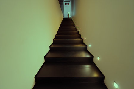 A long, modern staircase with dark wooden steps and built-in side lights. The staircase leads up to a hallway with a door at the top, illuminated by soft green and yellow lighting; wide-angle low-keyの写真素材