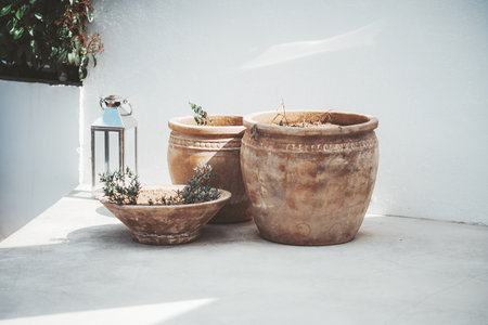 Three rustic clay pots with small plants are placed on a sunlit outdoor patio against a white wall. A metallic lantern sits beside, casting shadows and adding an elegant touch to a minimalist settingの写真素材