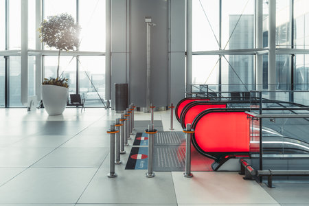 Modern airport interior with a red escalator, metal barriers, and a large potted plant under natural sunlight. The clean, spacious area features glass walls and a sleek, contemporary designの写真素材