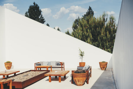 Modern outdoor patio with wooden furniture including benches and tables, arranged around a stone-filled fire pit. Terracotta planters with greenery and white walls; selective focus on a front clay potの写真素材