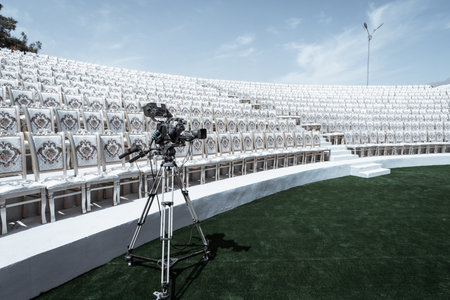 High-definition video camera on a tripod set up in an empty amphitheater with rows of ornate, white chairs. The scene features a clear blue sky, green artificial grass, and modern lighting fixturesの写真素材
