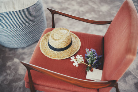 A straw hat with a black band, a small bouquet of purple and white flowers, and a blank envelope rest on a red upholstered chair. A blue-striped pouf is partially visible aloof on a carpeted floorの写真素材
