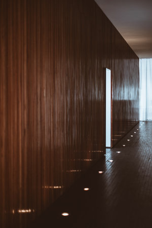 A sleek modern hallway with a wooden panel wall and floor, illuminated by recessed floor lights creating a warm ambiance. The hallway leads to a partially open door, revealing soft daylightの写真素材