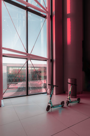 Two electric scooters are parked indoors next to a large floor-to-ceiling window in a modern, spacious building. The window reflects an exterior urban scene with a warm, pinkish lightの写真素材