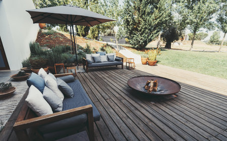 Outdoor patio area with wooden deck, cushioned sofas, and a fire pit. An umbrella provides shade, and the garden is landscaped with various plants and trees, creating a cozy ambianceの写真素材