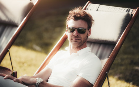 A caucasian man with curly brown hair and a white polo shirt is sitting in a deck chair outdoors on a sunny evening. He is wearing sunglasses and a wristwatch. A copy-space area on the leftの写真素材