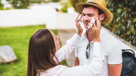 A woman applies sunscreen to a man's sunburned face. He wears a straw hat and sunglasses. The...