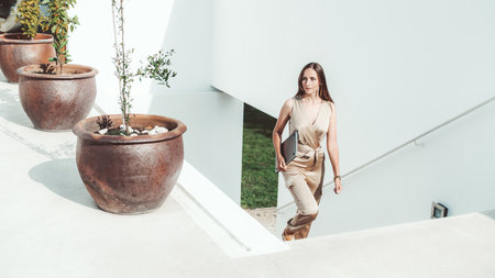 A woman in a beige jumpsuit holding a laptop walks up a white outdoor staircase lined with large terracotta pots containing small plants. Bright daylight illuminates the sceneの写真素材