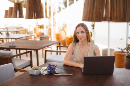 A hispanic businesswoman sits at an outdoor table with a laptop, a notebook, and a small flower arrangement. The setting features modern furniture, warm lighting, and decorative hanging lampsの写真素材
