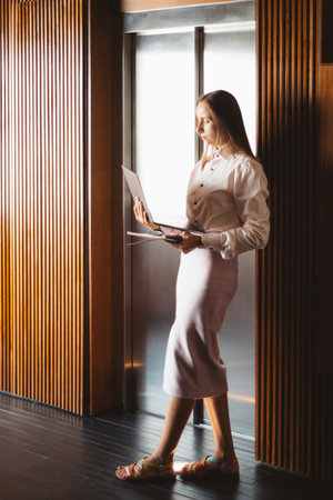 A businesswoman in a white shirt and pastel skirt stands leaning against an elevator door, working on her laptop. The interior features wooden paneling and soft lightingの写真素材