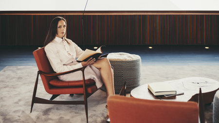 A hispanic woman is seated on an orange chair in a minimalist interior, holding an open book. The room is spacious with soft lighting, a modern design, and neutral tones - clean lines and simple decorの写真素材