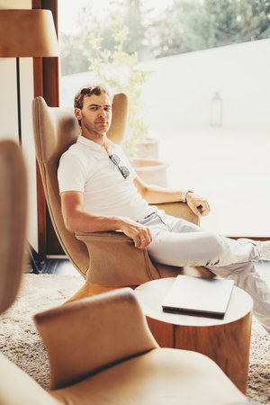 A man in a white shirt and pants sits casually in a modern, beige armchair, with his left arm resting on the chair's armrest and a laptop placed on a wooden table beside himの写真素材