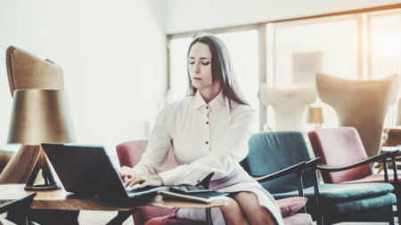 A professional hispanic woman entrepreneur in a beige dress stands against a plain white background, holding a laptop in her right hand, looking confidently into the camera, with a calm expressionの写真素材