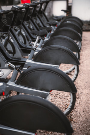 A close-up vertical shot with a shallow depth of field of aligned rental electric bicycles in a docking station on a gravel surface, focusing on wheels and saddles; uniformity and modern designの写真素材