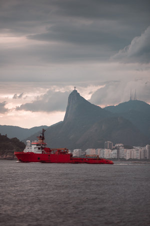 A vibrant red ship sails across the bay of Rio de Janeiro, with the iconic Christ the Redeemer statue on Corcovado Mountain in the background, under cloudy skies, near city buildingsの写真素材