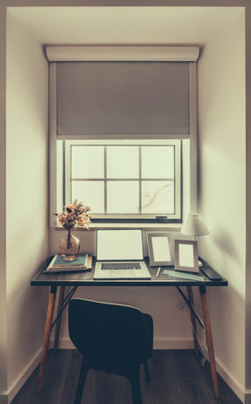 A minimalist workspace with a laptop, books, and photo frames on a modern black desk. A vase with dried flowers and a small lamp sit beside a window with a roller blind. Natural lightingの写真素材