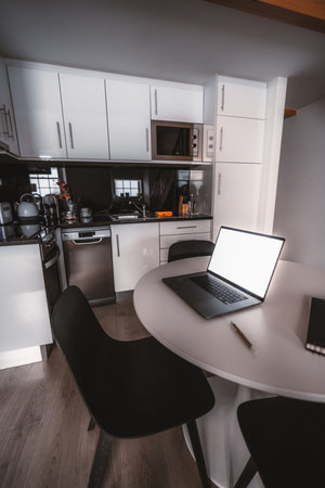A modern kitchen interior featuring white cabinets, stainless steel appliances including a microwave and dishwasher, a black round dining table, and a laptop on the tableの写真素材
