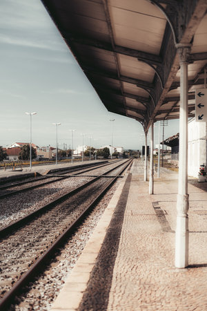 A vertical shot of a deserted train station platform with empty railway tracks extending into the distance. Covered shelter with metal support columns. Quiet urban surroundings under a clear skyの写真素材