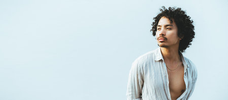 Portrait of a young asian man with curly hair, wearing an open, light linen shirt, looking thoughtfully to the side against a clean, soft blue background, conveying a serene moodの写真素材