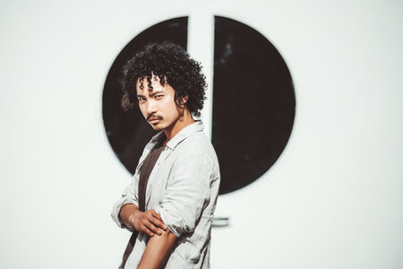 Portrait of a confident young asian man with curly hair standing against a modern black and white geometric background, wearing a light shirt over a dark t-shirt, looking at the cameraの写真素材