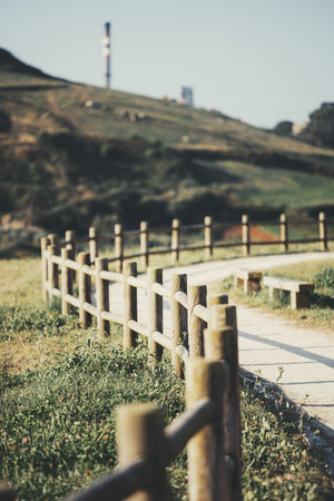 Wooden fence lines a winding dirt pathway, surrounded by green grass and wildflowers, leading up a grassy hillside. Industrial chimneys are visible in the blurred background; shallow depth of fieldの写真素材