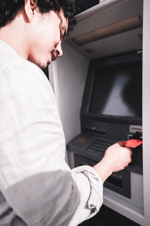 Man inserting a red bank card into an ATM, captured from a close side angle. The screen is dark, emphasizing the action of the user interacting with the cash machineの写真素材
