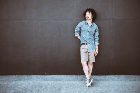 A stylish asian man standing against a dark wall, dressed in a denim shirt, beige shorts, and...