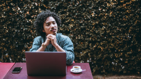 Young asian man with curly hair in a thoughtful pose, seated outdoors with a laptop, phone, and...