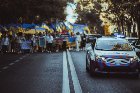 Ukrainian anti-war protest, a defocused crowd holding national flags and anti-war signs in a public square. Demonstrators showing strong resistance; selective focus on a police car on the rightの写真素材